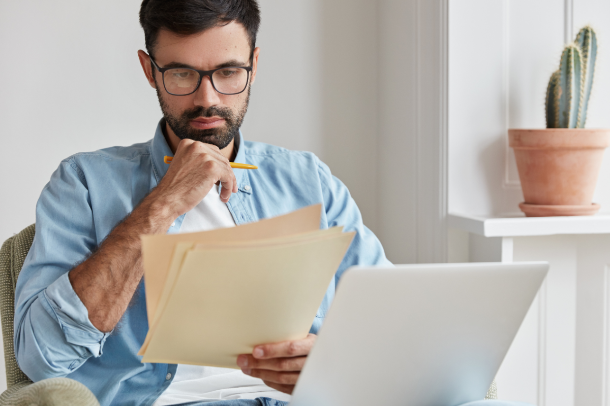 Homem em ambiente de escritório sentado em frente a um notebook lendo documentos em mãos representando: Como ficará o crédito de imposto na Reforma Tributária?