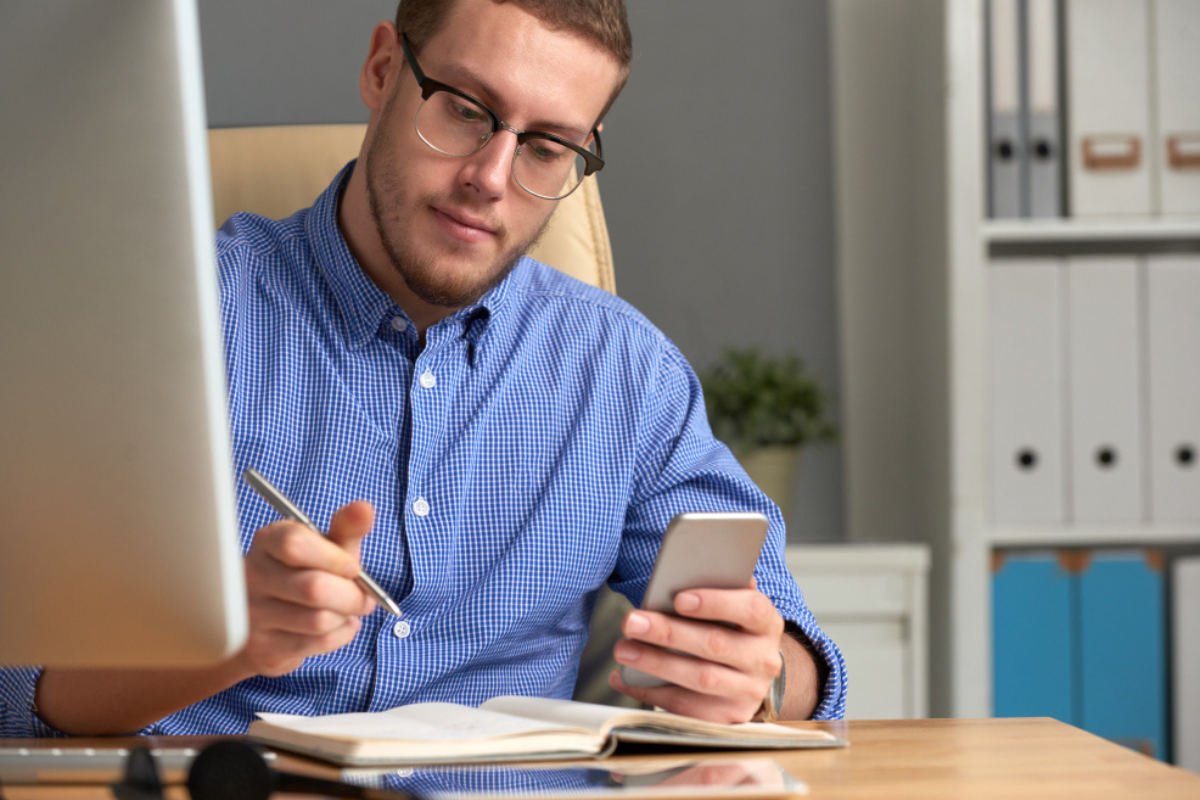 Homem sentado fazendo anotações em uma caderneta sobre uma mesa e usando celular em ambiente de casa representando: Imposto Seletivo: qual o motivo da sua criação?