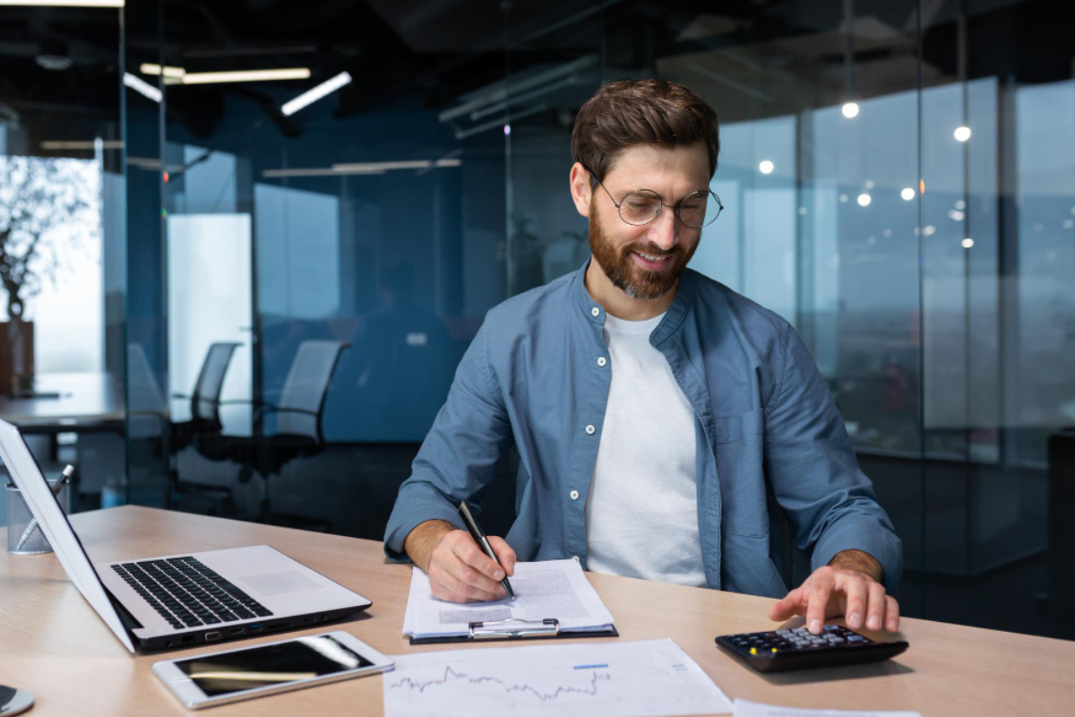 Homem sentado em frente a um notebook sobre uma mesa fazendo anotações em uma prancheta e usando uma calculadora e ambiente de escritório representando: Imposto sobre Valor Agregado (IVA): o que é e o que muda com ele.