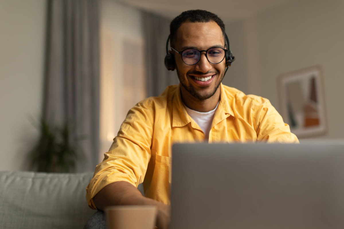 Homem sentado usando notebook com fone de ouvido em ambiente de sala de estar representando: Como ficam as alíquotas estaduais e municipais com o IBS?