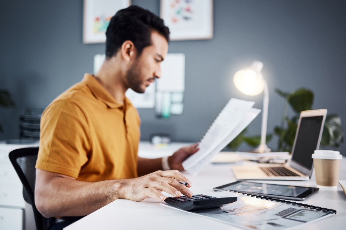 Homem em ambiente de escritório sentado em frente a um computador com documentos na mão esquerda e usando uma calculadora sobre uma mesa com a mão direta representando: IBS: entenda o substituto do ICMS e ISS.