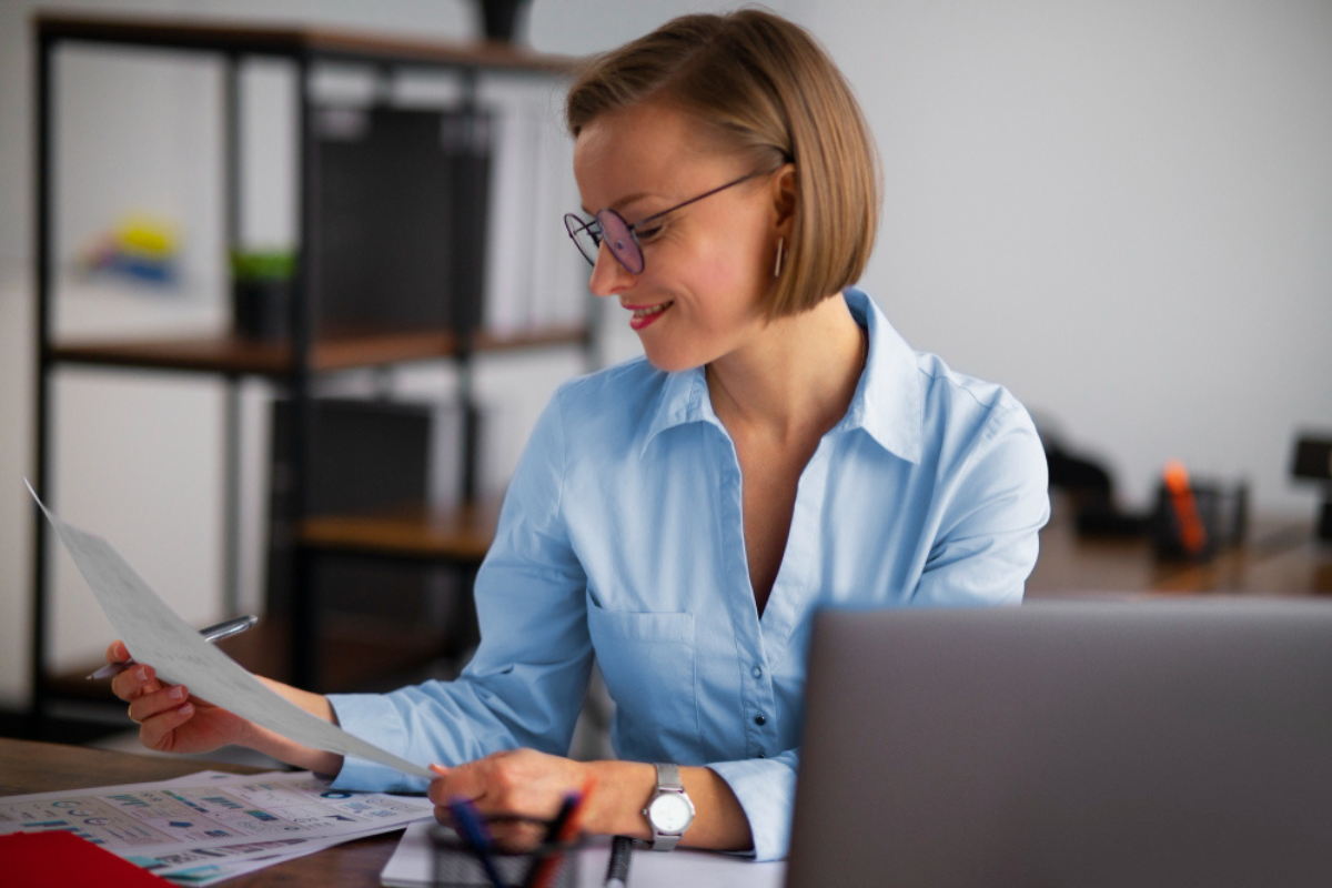 Mulher em ambiente de escritório sentada em frente a um computador fazendo leitura de documentos em mãos representando: Extinção do PIS/Cofins: como a CBS os substituirá?