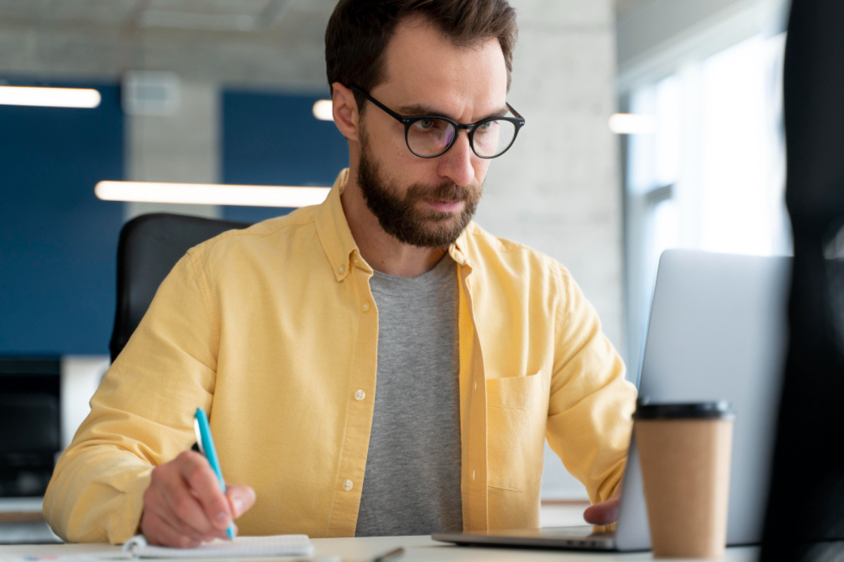 Homem sentado em frente a um notebook sobre uma mesa com copo ao lado e fazendo anotações em ambiente de escritório representando: Imposto Seletivo (IS): quais são os setores e produtos afetados?