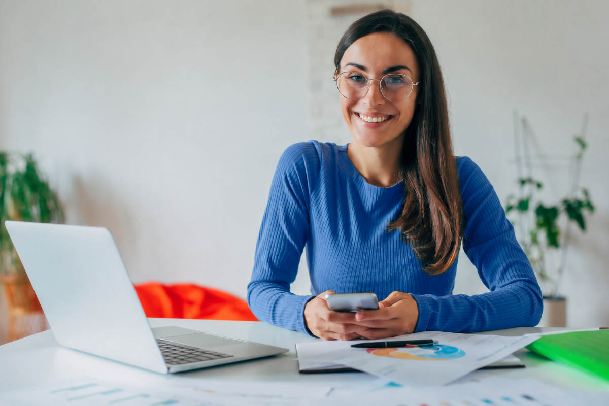 Moça trabalhando em um notebook sobre uma mesa e com celular em mãos em ambiente de escritório representando: Rejeição 232: IE do destinatário não informada.