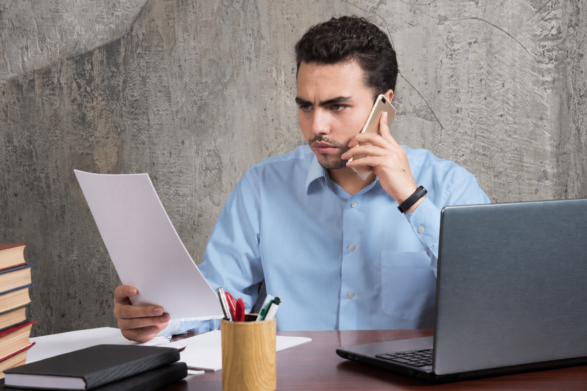 Profissional falando ao celular lendo um documento sentado em frente ao notebook sobre uma mesa em ambiente de escritório representando: O que acontece se sua empresa parar de emitir notas fiscais? Entenda os riscos.