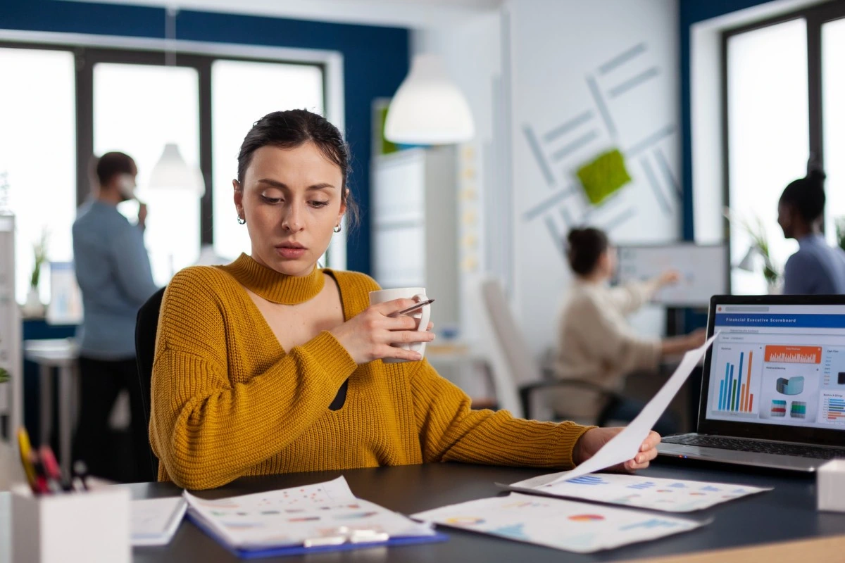 Mulher sentada trabalhando em seu notebook sobre uma mesa e analisando documentos e relatórios sobre o que é ponto de equilíbrio e como calculá-lo, em ambiente de escritório com colegas ao fundo.