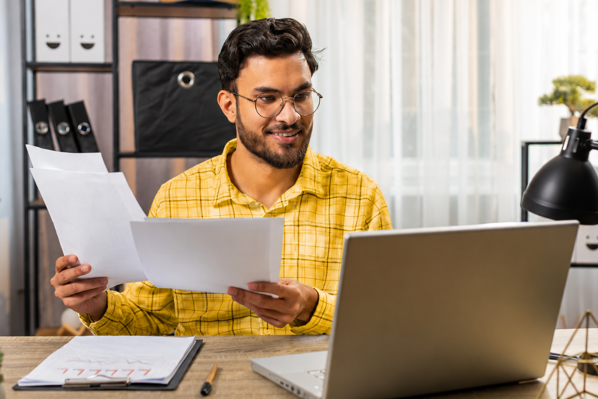 Homem analisando documentos em mãos e trabalhando em seu notebook em ambiente de escritório representando: Por que o estoque errado impacta imposto?