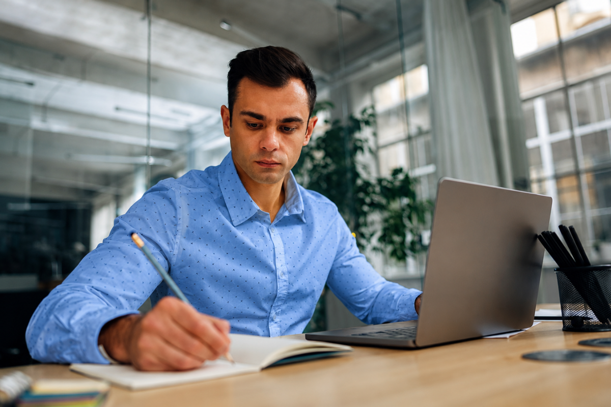 Homem sentando a mesa com notebook escrevendo em um papel representando: Elisão fiscal e NFC-e para varejistas no Simples Nacional: o que a lei permite e onde estão os limites